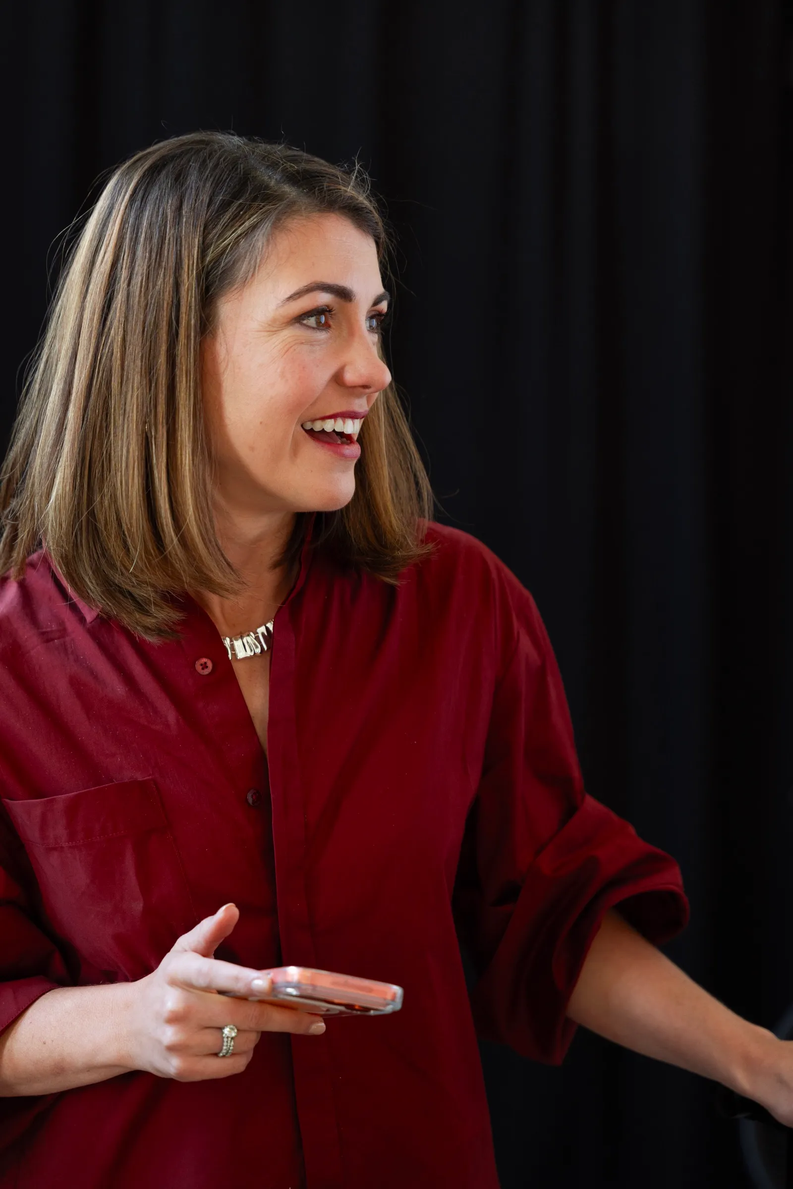 A woman in a red shirt laughs, phone in hand, looking off to one side